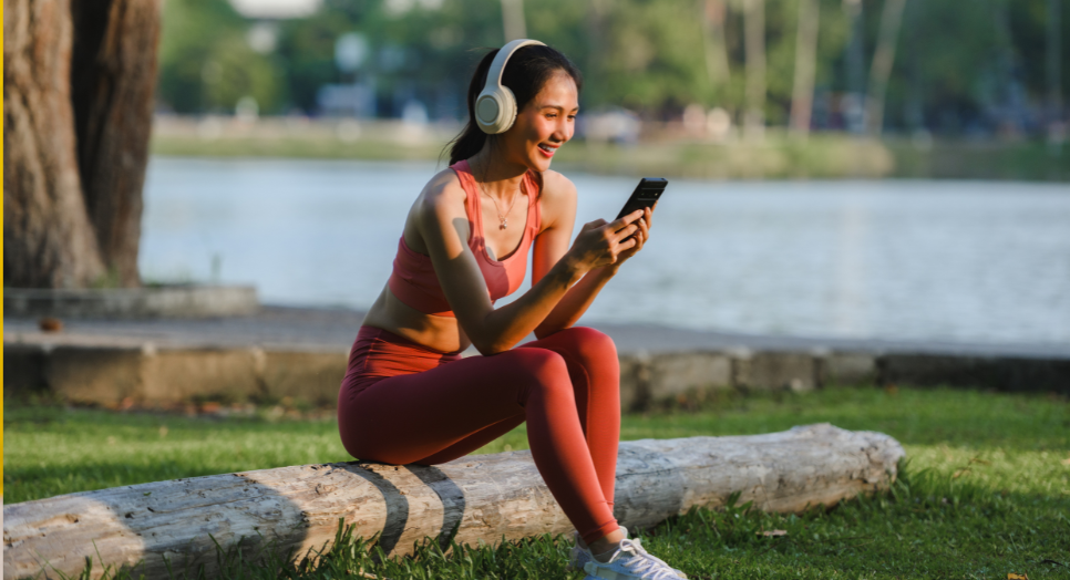 A woman sitting on a log outside looking at her phone while wearing headphones and workout clothes 