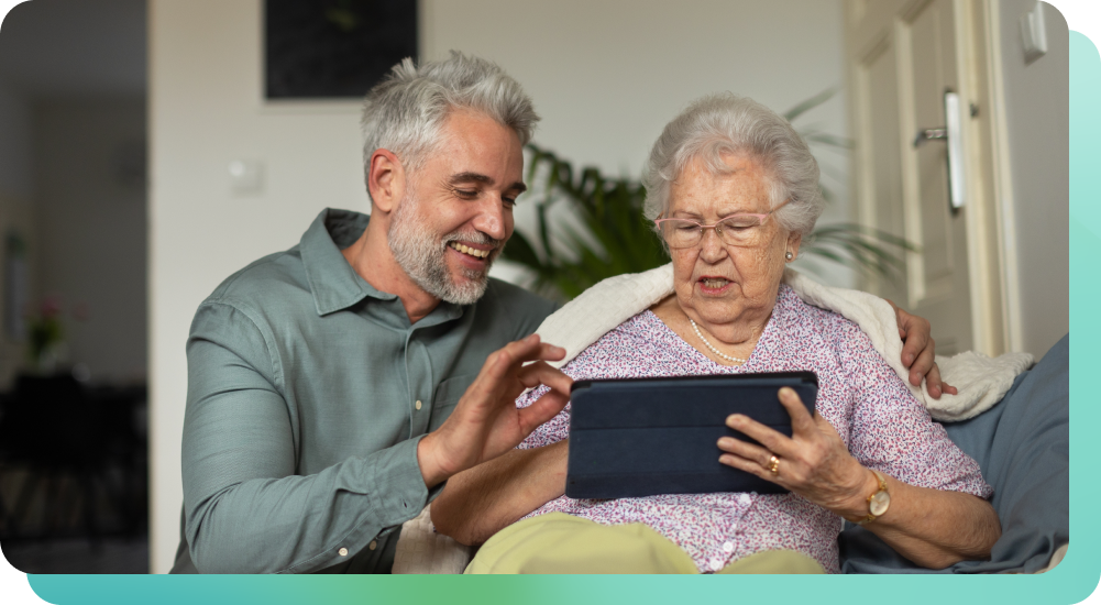 Man guiding an elderly woman on a couch as she explores music for wellness on a tablet, both smiling