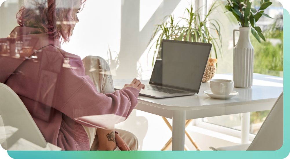Woman in pink sweatshirt at a sunlit table typing on a laptop, using functional music for focus