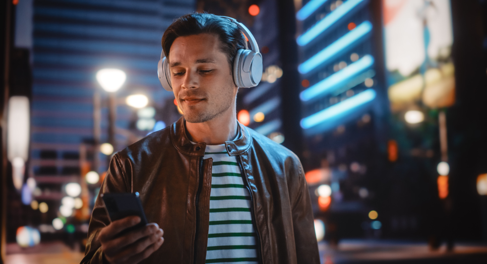 Man wearing headphones and holding a phone while walking in a city at night, listening to relaxing summer releases from Feed Originals.-1