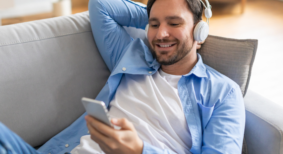 Smiling man relaxing on a couch wearing white headphones and looking at his smartphone, enjoying music in a cozy living room setting.