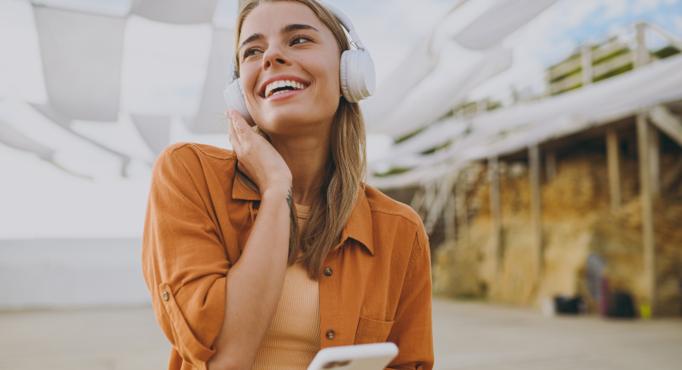 Smiling woman listening to music on wireless headphones while holding a smartphone outdoors, wearing a rust-colored shirt and crop top under a sunny sky.