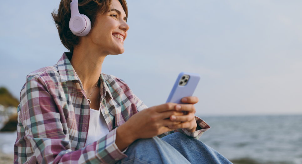 Smiling woman wearing headphones and holding a smartphone while listening to music by the beach