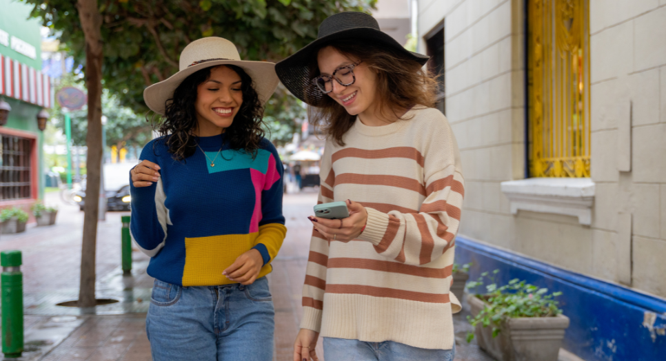 Two young women wearing hats and casual sweaters walking down a city street, smiling and looking at a smartphone together.