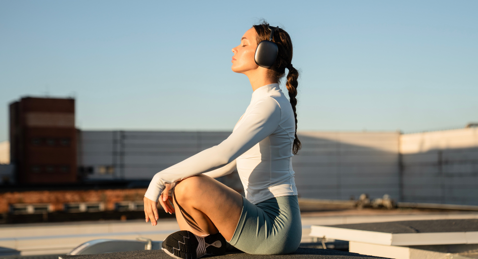 Woman in athletic wear sitting cross-legged outdoors with headphones on, eyes closed, meditating while listening to music for focus and relaxation.