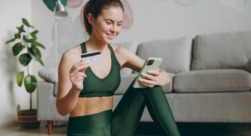 Woman in green activewear sitting on a yoga mat, smiling while holding a smartphone and a credit card, suggesting an online fitness app purchase or HSA payment.