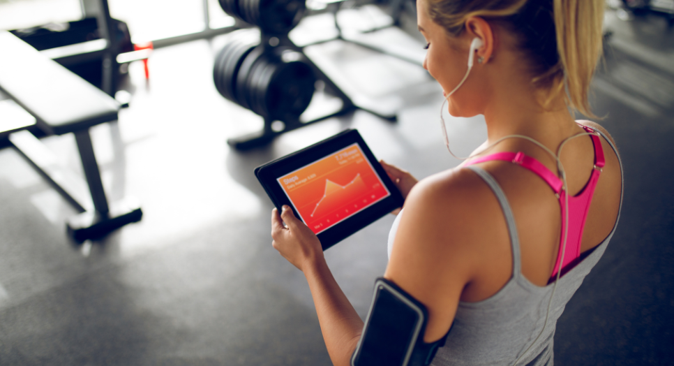 Woman in gym using a tablet to track fitness progress, wearing headphones and an armband device